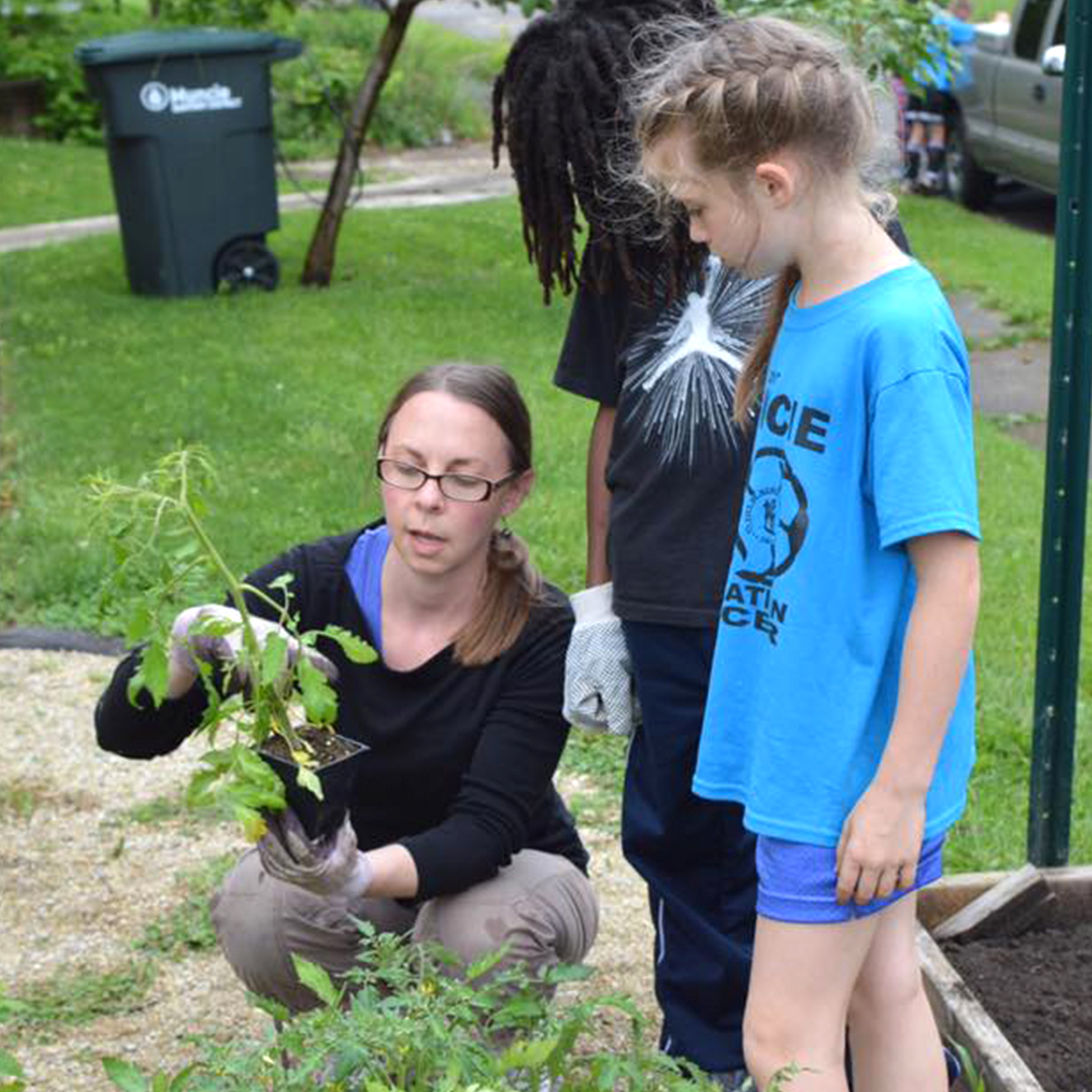 Image of a woman squatting down and talking to explain a plant to younger children in the community garden.