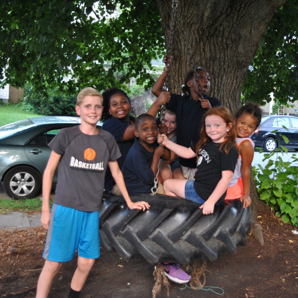 Seven children crowded onto a tire swing, all looking at the camera and smiling.