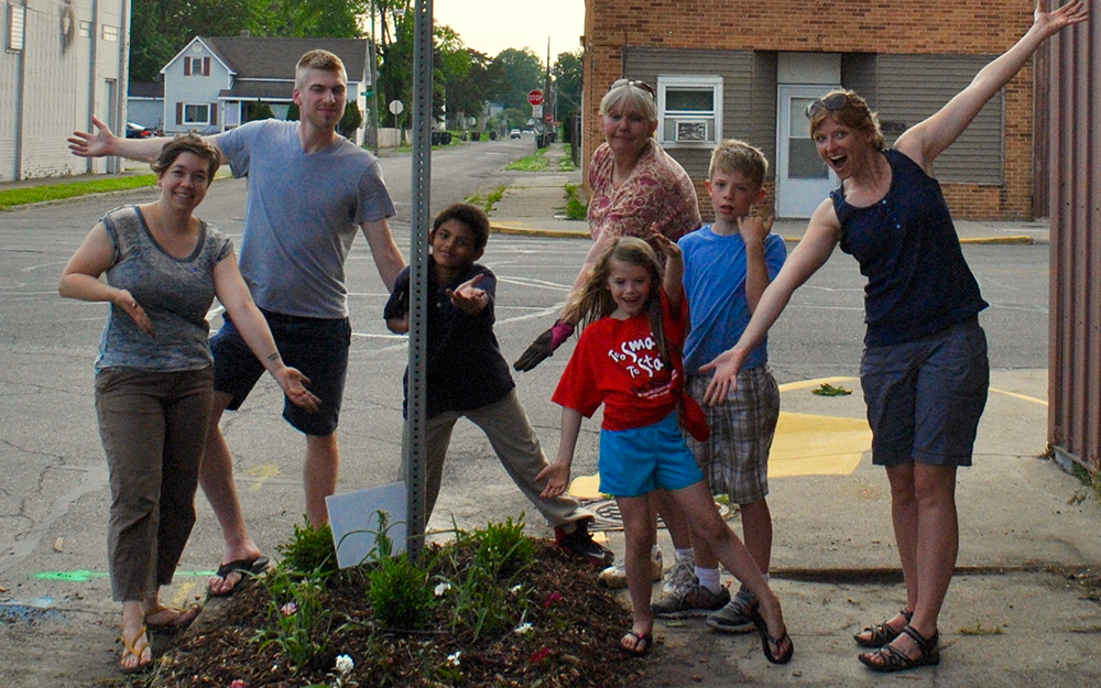 Three young children and four adults in varying arms-outstretched poses celebrating their mini-garden at the base of a stop sign.